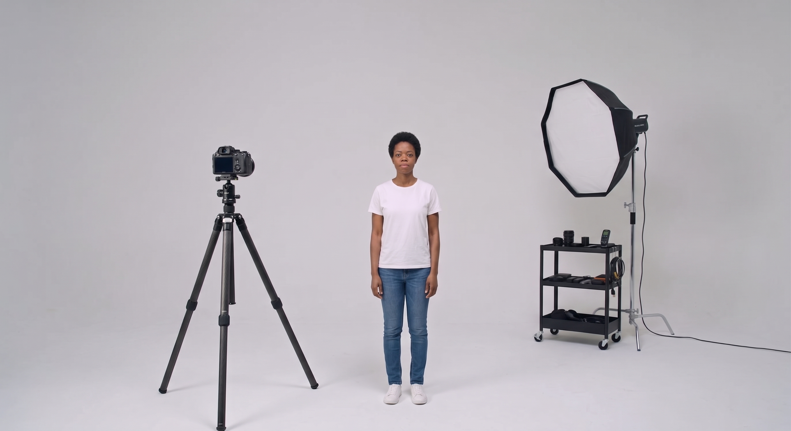 Tipos de planos: Plano Geral. Uma mulher centralizada, usando camiseta branca e calças jeans, com objetos de estúdio fotográfico ao redor dela