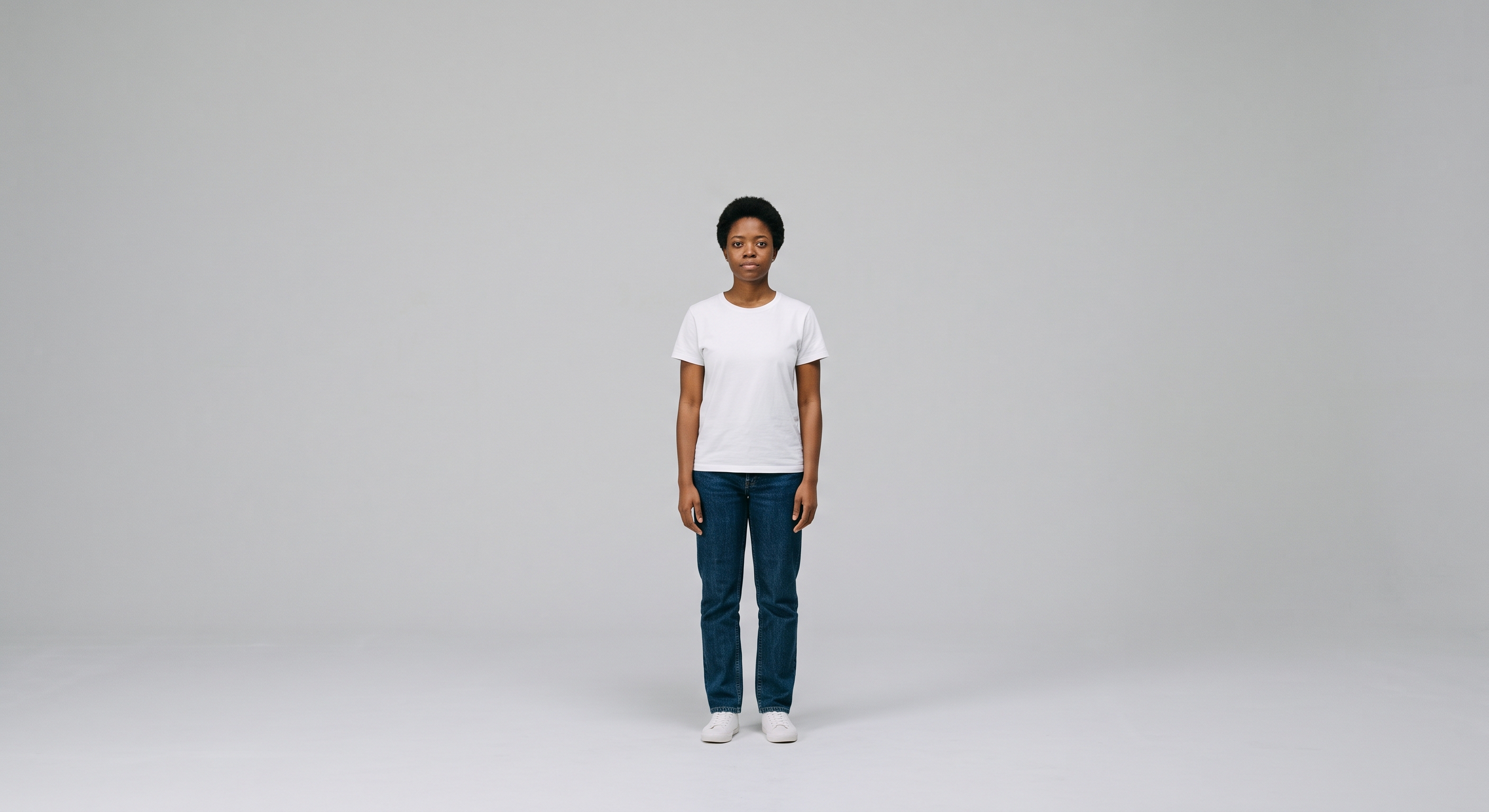 Tipos de plano: plano aberto. Uma mulher ao centro da imagem, usando calça jeans e camiseta branca em plano aberto com um fundo neutro.